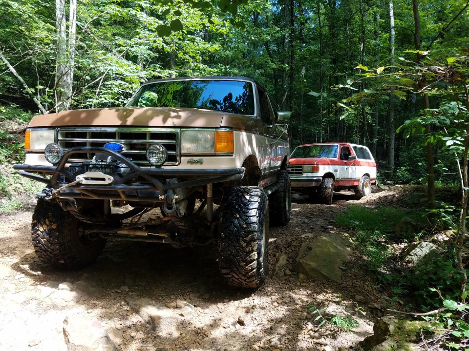 Brown and tan lifted SUV with winch, red and white SUV behind on forest trail