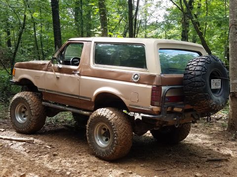 1988 Ford Bronco - This lifted 1988 Ford Bronco, seen off-road, features a two-tone brown paint scheme and a rear-mounted spare tire.