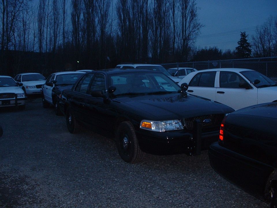 Black Ford Crown Victoria on steel wheels at dusk in a fenced lot of light-colored cars