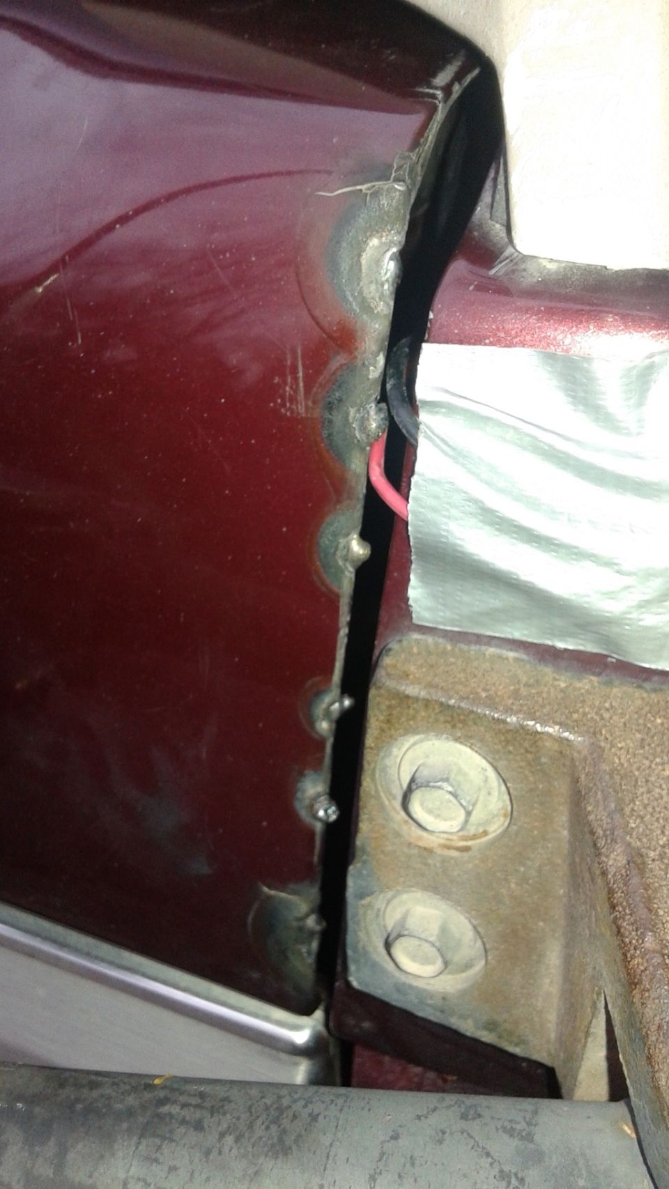 Close-up of a dark red car door seam showing crude welds, exposed wiring, and silver tape