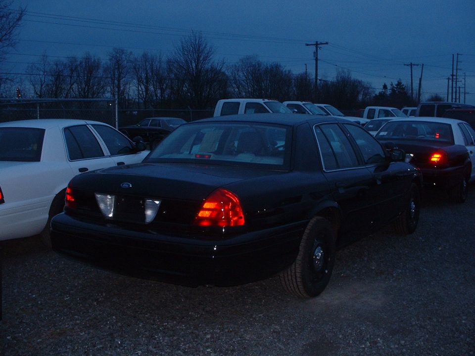 Rear of black Ford Crown Victoria at dusk with taillights lit among parked sedans