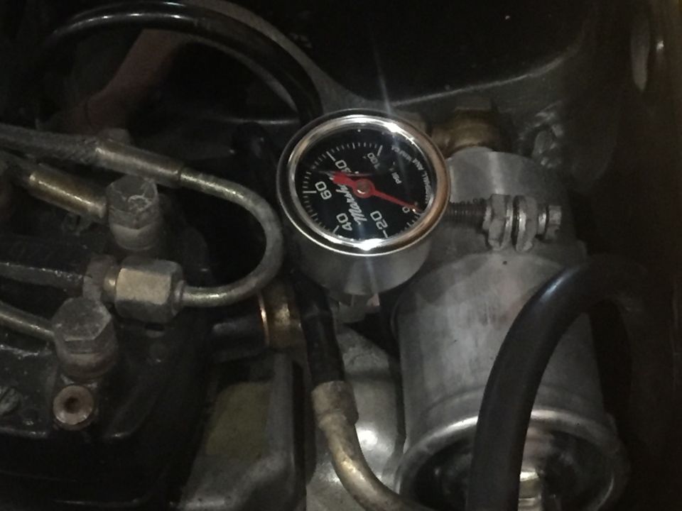 Close-up of a fuel pressure gauge with red needle pointing to about 25 PSI, surrounded by engine components and fuel lines