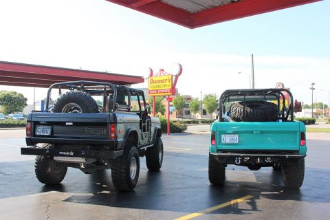1972 Ford Bronco - Two Ford Broncos, a gray and a teal model, are parked side-by-side outside a restaurant.