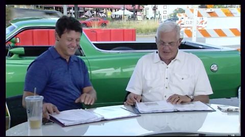 1971 Ford Ranchero - Two men sit at a table in front of a green Ford Ranchero. The man on the left wears a blue polo shirt and smiles while looking down at pape…