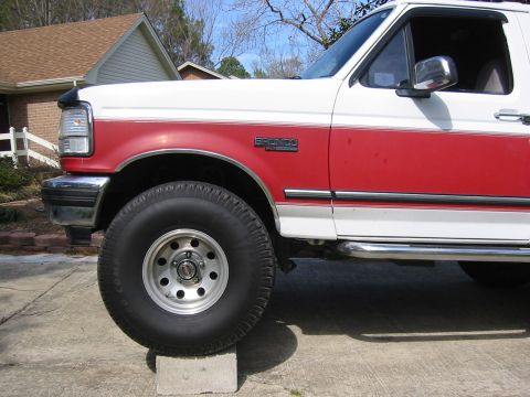 1993 Ford Bronco - The front quarter panel and large tire of a white and red 1993 Ford Bronco are visible, with the vehicle resting on a concrete block.