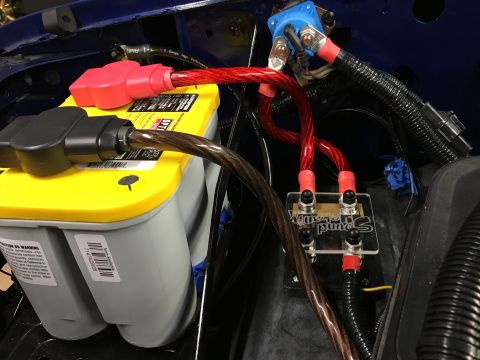 1996 Ford Bronco - A close-up view of a car battery and its wiring setup, featuring red and black cables connected to a distribution block. The battery is a y…