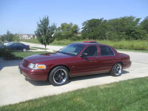 2004 Ford Crown Victoria - A dark red 2004 Ford Crown Victoria is parked on a concrete driveway with a grassy lawn in the foreground and a line of trees and houses in…
