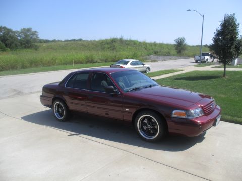 2004 Ford Crown Victoria - A maroon 2004 Ford Crown Victoria sedan is parked on a concrete driveway on a sunny day. The car has aftermarket black wheels with chrome t…
