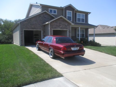 2004 Ford Crown Victoria - A maroon Ford Crown Victoria sedan is parked in a concrete driveway in front of a two-story house with a garage. The car's rear is facing t…