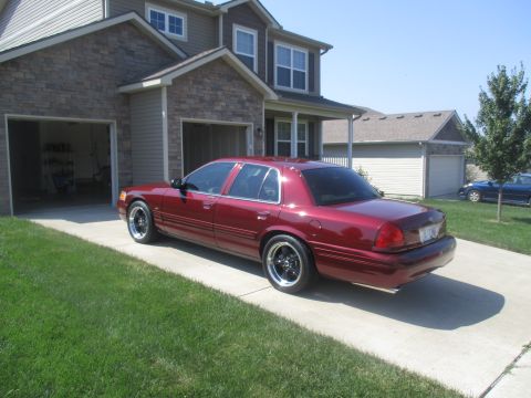 2004 Ford Crown Victoria - A dark red Ford Crown Victoria sedan is parked on a concrete driveway in front of a residential house with two garage doors. The car has af…