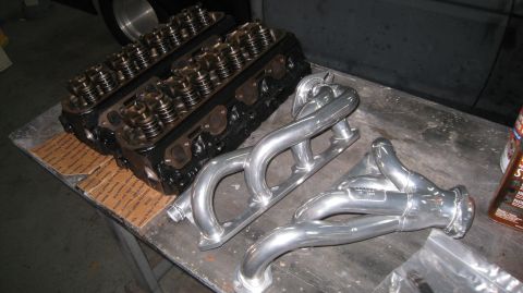 1996 Ford Bronco - Two black engine cylinder heads with visible valve springs and rocker arms sit on a table next to two polished aluminum exhaust headers.
