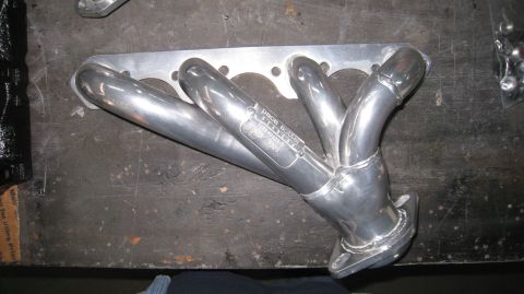 1996 Ford Bronco - A polished stainless steel exhaust header with four pipes merging into one is laid on a dark, textured surface. A label on one of the pipes…