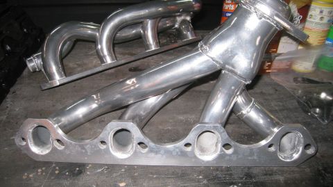1996 Ford Bronco - A polished aluminum exhaust manifold is shown on a workbench. The manifold has four pipes that merge into a single outlet, with a flange at…