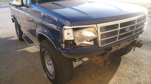 1996 Ford Bronco - The front driver's side of a dark blue Ford Bronco is shown, with a damaged headlight assembly and a rusty front bumper. The vehicle is par…