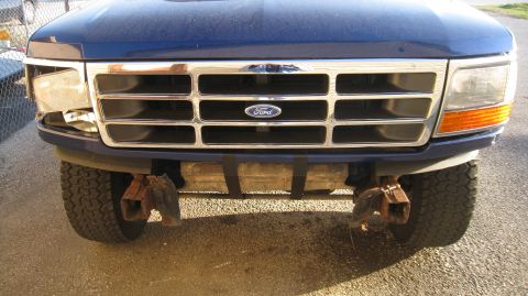 1996 Ford Bronco - The front view of a blue Ford Bronco with a damaged headlight and a chrome grille. A rectangular object is strapped to the front bumper.