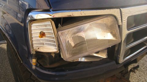 1996 Ford Bronco - Close-up of the damaged front passenger side headlight and turn signal assembly on a dark blue vehicle. The headlight lens is cracked and d…