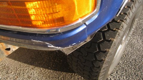 1996 Ford Bronco - Close-up view of the front corner of a blue vehicle, showing the orange turn signal, a damaged blue bumper, and a tire. The vehicle appears…