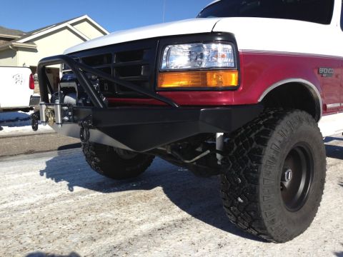 1996 Ford Bronco - A close-up, low-angle shot of the front of a white and red Ford Bronco with an aftermarket black steel bumper and winch. The vehicle is par…
