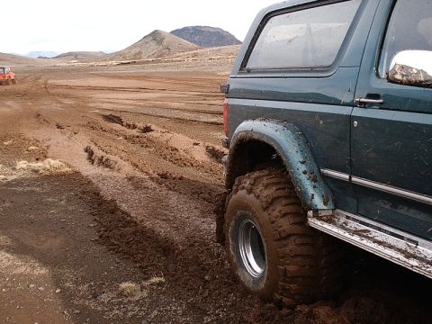 1996 Ford Bronco - A 1996 Ford Bronco navigates a muddy off-road trail, with another vehicle visible in the distance.