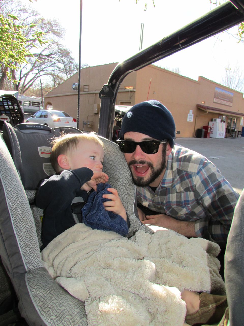Man in sunglasses and plaid shirt smiling next to a child in a car seat.