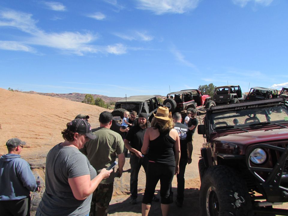 People gathered around off-road Jeeps on a sandy trail, with a red Jeep Wrangler in the foreground.