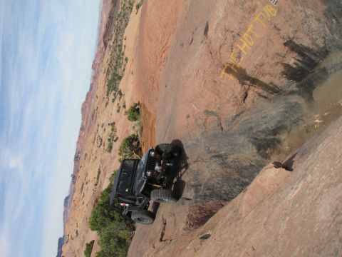 1995 Jeep Wrangler - A black Jeep YJ is navigating a steep, rocky incline in a desert landscape. Yellow text painted on the rock face reads "THE HOT TUB".
