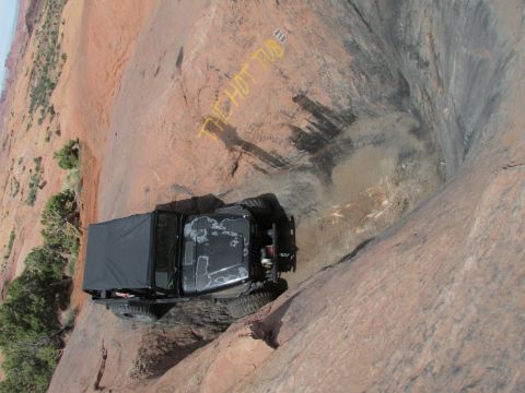 1995 Jeep Wrangler - A black Jeep YJ with a soft top is parked on a steep, rocky, desert landscape. Yellow text reading "THE HOT TUB" is visible on the rock fac…
