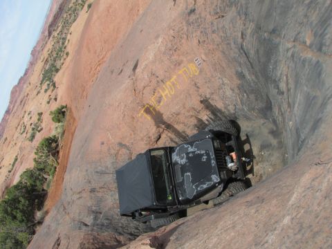 1995 Jeep Wrangler - A black Jeep YJ with a soft top is positioned precariously on a steep, rocky incline. Yellow spray paint on the rock reads "THE HOT TUB".