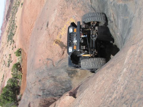 1995 Jeep Wrangler - A black Jeep YJ is tilted precariously on its side, with its tires and undercarriage visible, navigating a steep, rocky canyon. Yellow spra…