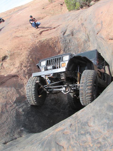 1995 Jeep Wrangler - A black Jeep Wrangler YJ is navigating a steep, rocky off-road obstacle. A person is seated on the rocks above, watching the vehicle.