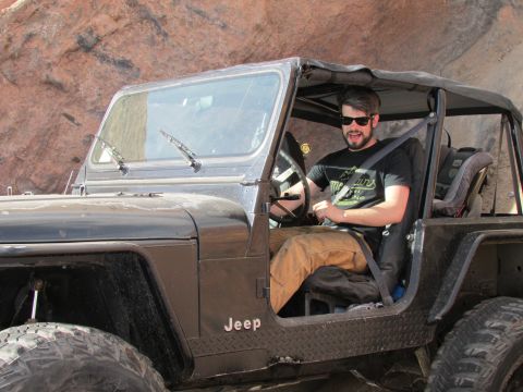 1995 Jeep Wrangler - A man wearing sunglasses and a black t-shirt is driving a black Jeep YJ with the top down. The vehicle is on an off-road trail with rocky t…