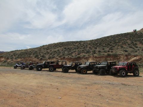 1995 Jeep Wrangler - A line of off-road vehicles, including several Jeep Wranglers and two UTVs, are parked on a dirt road in front of a scrub-covered hillside.