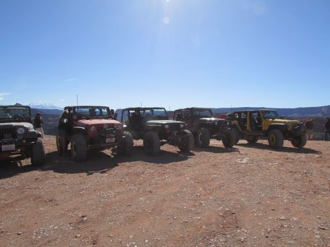 1995 Jeep Wrangler - A line of five Jeep Wranglers are parked on a dirt road with a bright blue sky overhead. The Jeeps are mostly red, yellow, and dark gray, w…