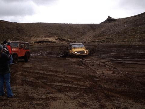 1996 Ford Bronco - Two off-road vehicles navigate a muddy terrain under an overcast sky, with onlookers observing the action.