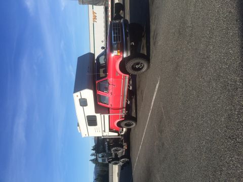 1995 Jeep Wrangler - A red pickup truck with a camper shell attached is parked on asphalt. The truck has large off-road tires and a lifted suspension.