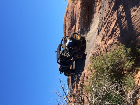 1995 Jeep Wrangler - A modified Jeep YJ is angled precariously on a steep, rocky incline with a bright blue sky in the background.