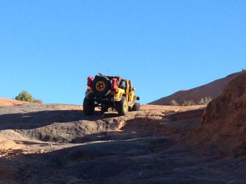 1995 Jeep Wrangler - A yellow Jeep Wrangler is driving up a rocky, off-road trail under a clear blue sky. The Jeep has a spare tire mounted on the back, along w…