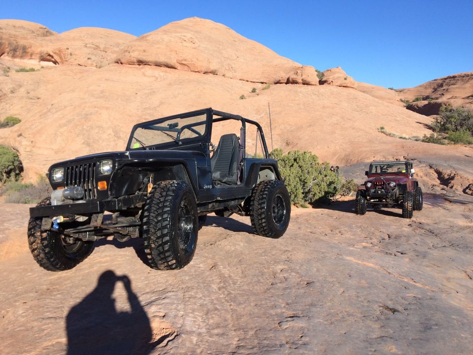 Black Jeep YJ parked on a rocky desert trail with a red Jeep Wrangler in the background.