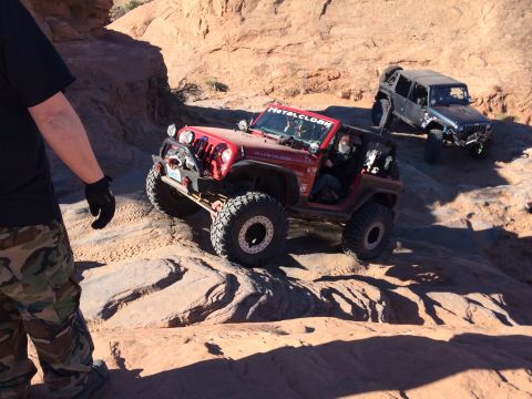 1995 Jeep Wrangler - A red Jeep Wrangler is navigating a rocky, off-road trail, with a black Jeep Wrangler following behind. The red Jeep has a winch and auxili…