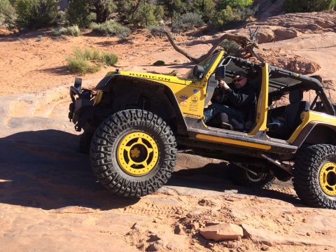 1995 Jeep Wrangler - A yellow Jeep Wrangler Rubicon is navigating a rocky, desert terrain. The driver is visible behind the wheel, wearing sunglasses and a hat.