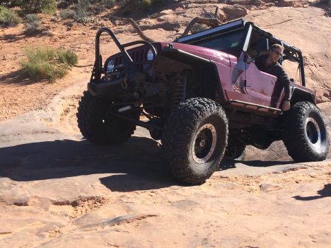 1995 Jeep Wrangler - A maroon Jeep YJ with large off-road tires is navigating a rocky, desert terrain. A person is visible in the driver's seat, smiling.
