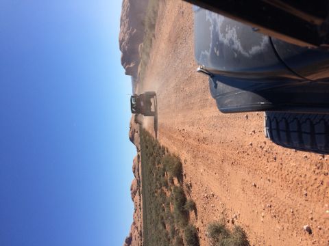 1995 Jeep Wrangler - A black Jeep YJ is captured from a low angle, driving on a dusty dirt road with red rock formations and sparse vegetation on the side. The…