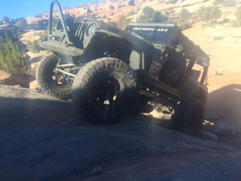 1995 Jeep Wrangler - A heavily modified olive green Jeep YJ is shown from a low angle, navigating a rocky, off-road terrain. The vehicle features large off-road…