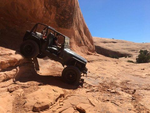 1995 Jeep Wrangler - A dark-colored Jeep YJ is navigating a steep, rocky, orange desert terrain under a clear blue sky. The vehicle is angled on the incline, wi…