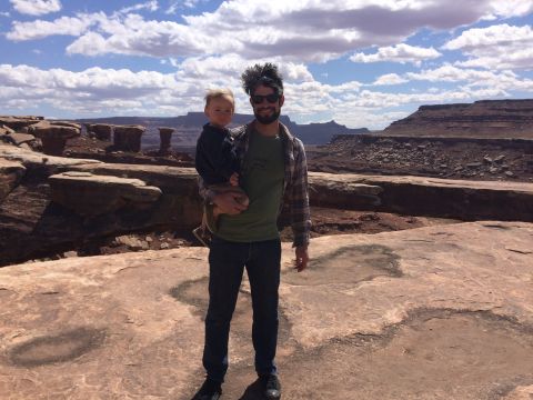 1995 Jeep Wrangler - A man holds a young child while standing in a desert landscape with rock formations and a cloudy sky.