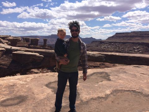 1995 Jeep Wrangler - A man holds a young child while standing in a desert landscape with rock formations and a cloudy blue sky.