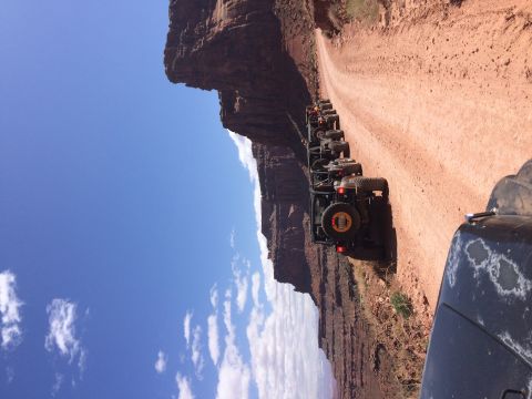 1995 Jeep Wrangler - A line of off-road vehicles, including a Jeep YJ, drive down a dusty dirt road alongside a towering red rock canyon under a bright blue sky…