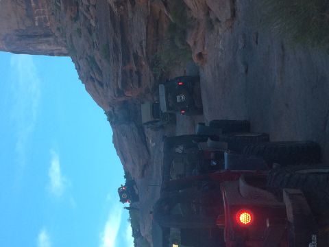 1995 Jeep Wrangler - A line of off-road vehicles, including Jeeps, are parked on a dirt path next to a large rock formation. The sky is blue with scattered clou…