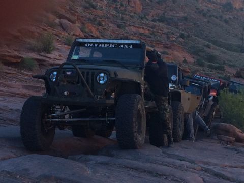 1995 Jeep Wrangler - A line of modified Jeeps are parked on a rocky, uneven terrain with a steep, vegetated hillside in the background. The lead Jeep has a ligh…