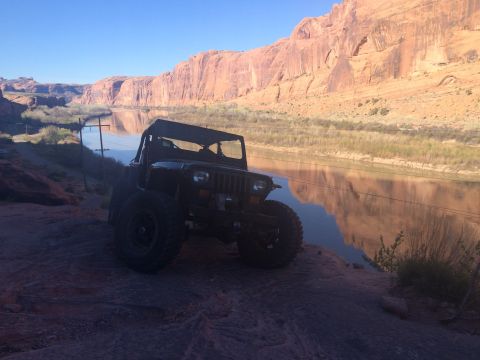 1995 Jeep Wrangler - A dark-colored Jeep YJ is parked on a rocky outcrop overlooking a wide river with red rock cliffs in the background.